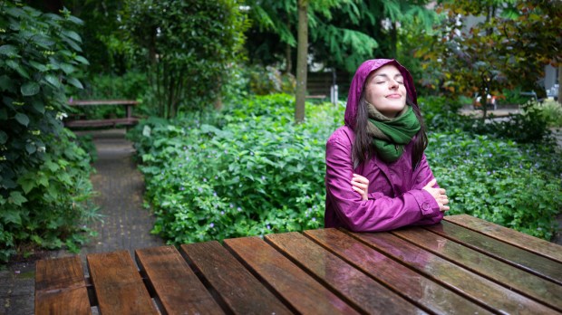 Beautiful young woman enjoying the rain in a garden decorated with eyes closed sits wooden table