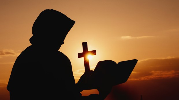 A monk in a hood with a crucifix in his hands stands against the backdrop of a dramatic sky at sunset