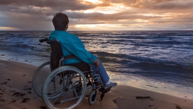 Social theme A disable woman in a wheelchair looks at a sunset on the beach by the sea Her folding wheelchair is part of her life serving as her legs Evening Dark key